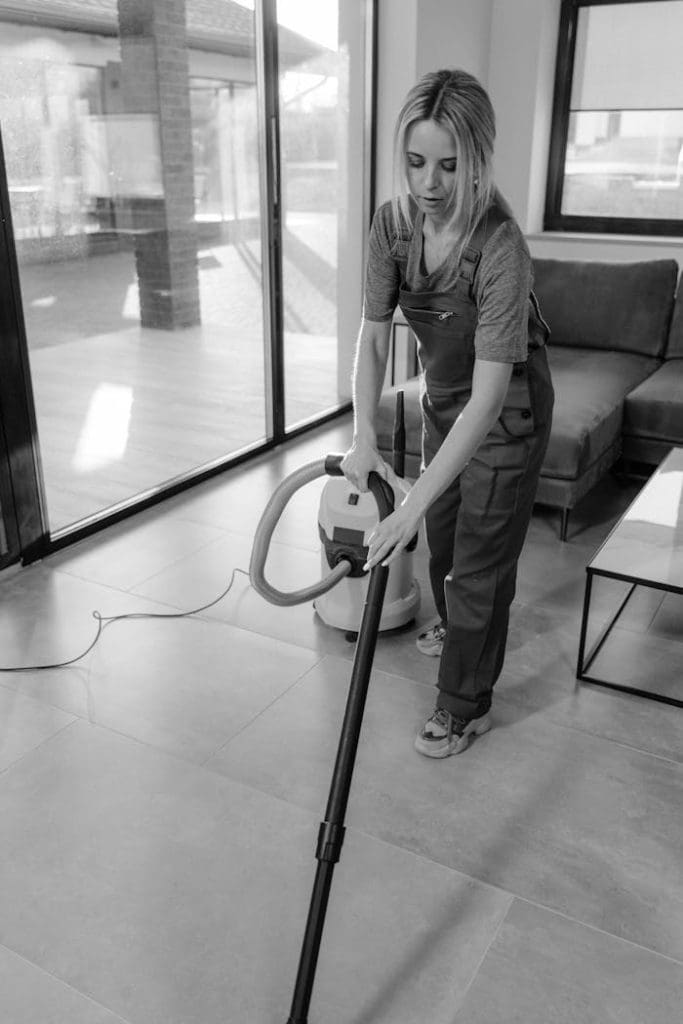 a-woman-cleaning-inside-a-house-6195287 A woman in work clothes vacuuming indoors, emphasizing cleanliness and efficiency.